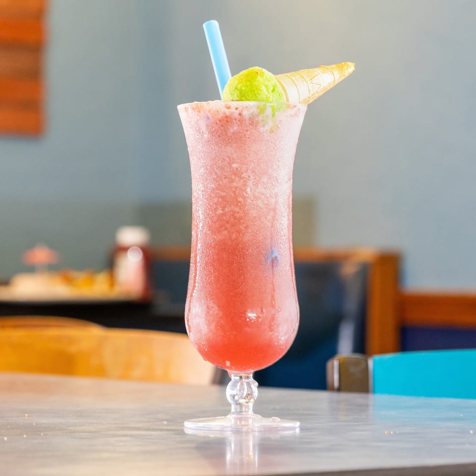Pink frozen drink in a tall glass garnished with a green scoop, waffle cone, and blue straw on a restaurant table.