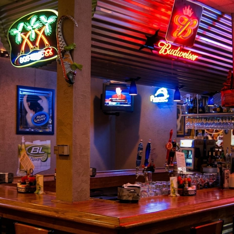 A lively bar interior with neon signs, multiple TVs, and a wooden counter with stools.