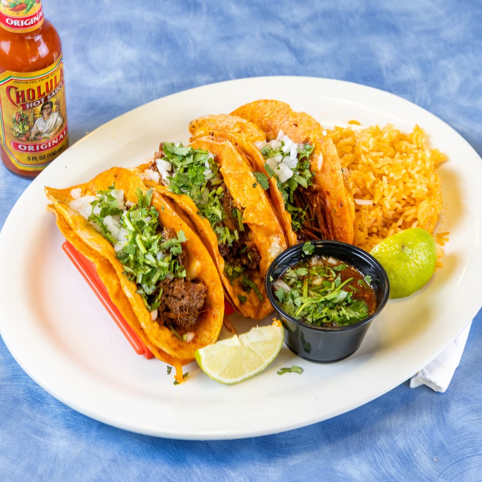 Plate of orange birria tacos topped with cilantro and onions, served with rice, consommé, lime, and hot sauce.