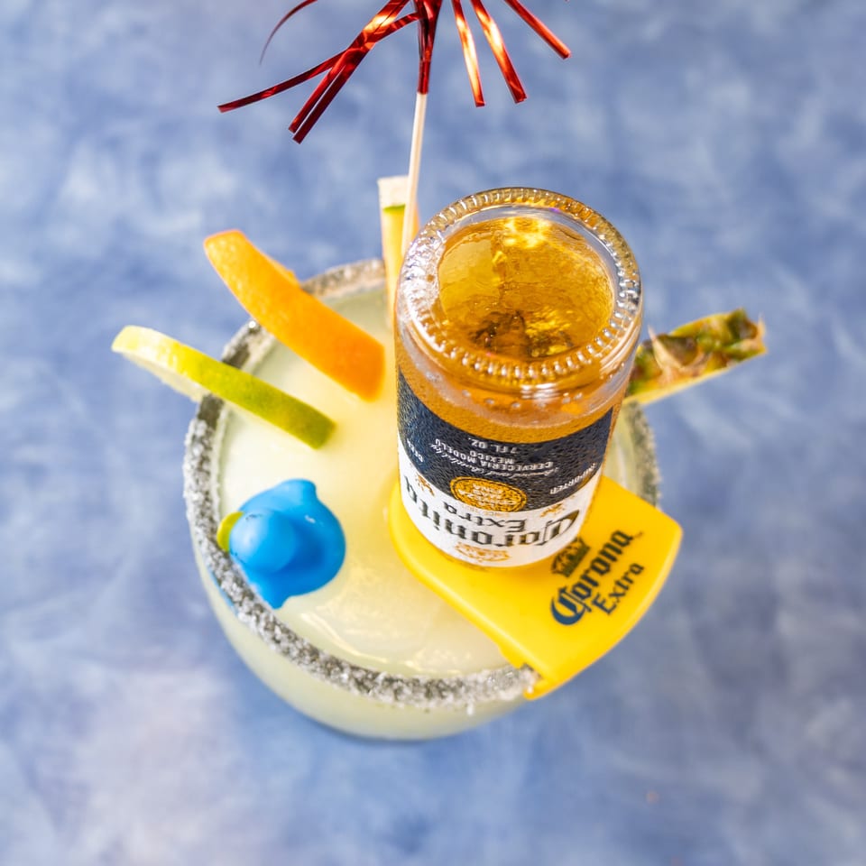 Margarita with a salted rim, lime and orange garnishes, blue candy, and a miniature Corona Extra bottle under a red cocktail umbrella.