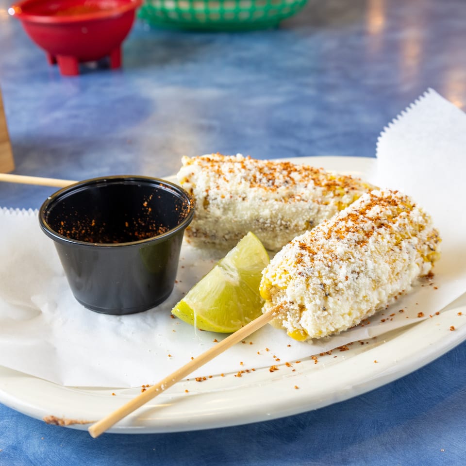 Two servings of Mexican street corn coated with cheese and spices, accompanied by lime wedges and dipping sauce on a plate.