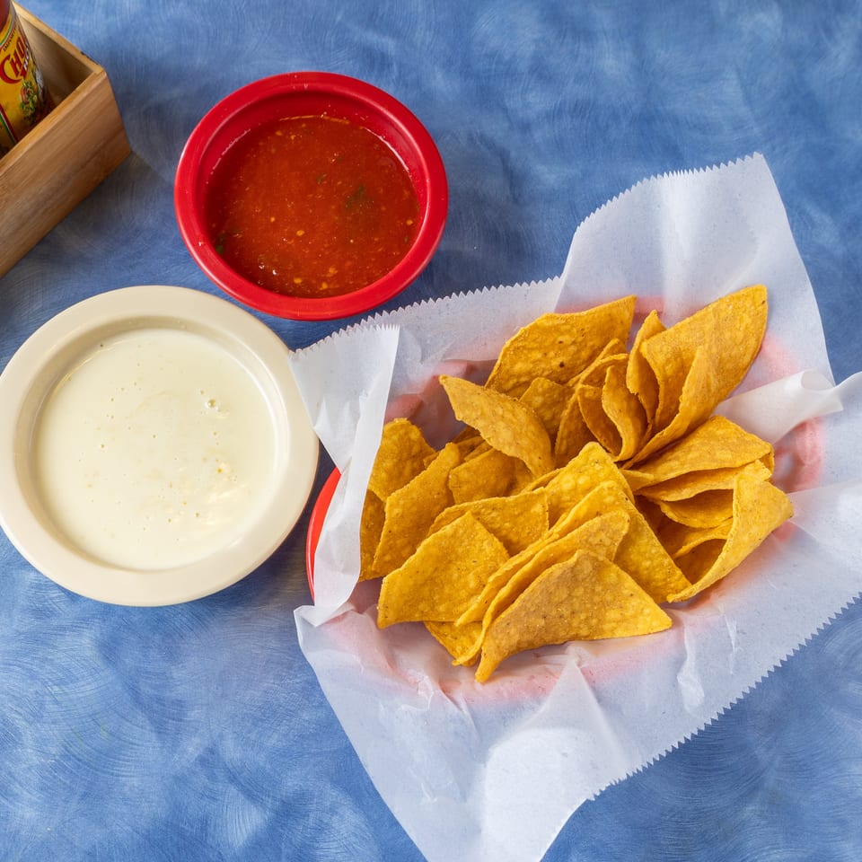 Basket of tortilla chips with red salsa and creamy white queso beside tabletop salt and hot sauce.