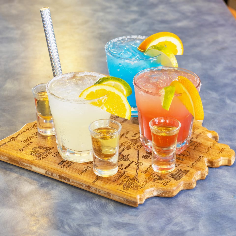 Colorful cocktails and shot glasses with citrus garnishes arranged on an engraved wooden serving board.