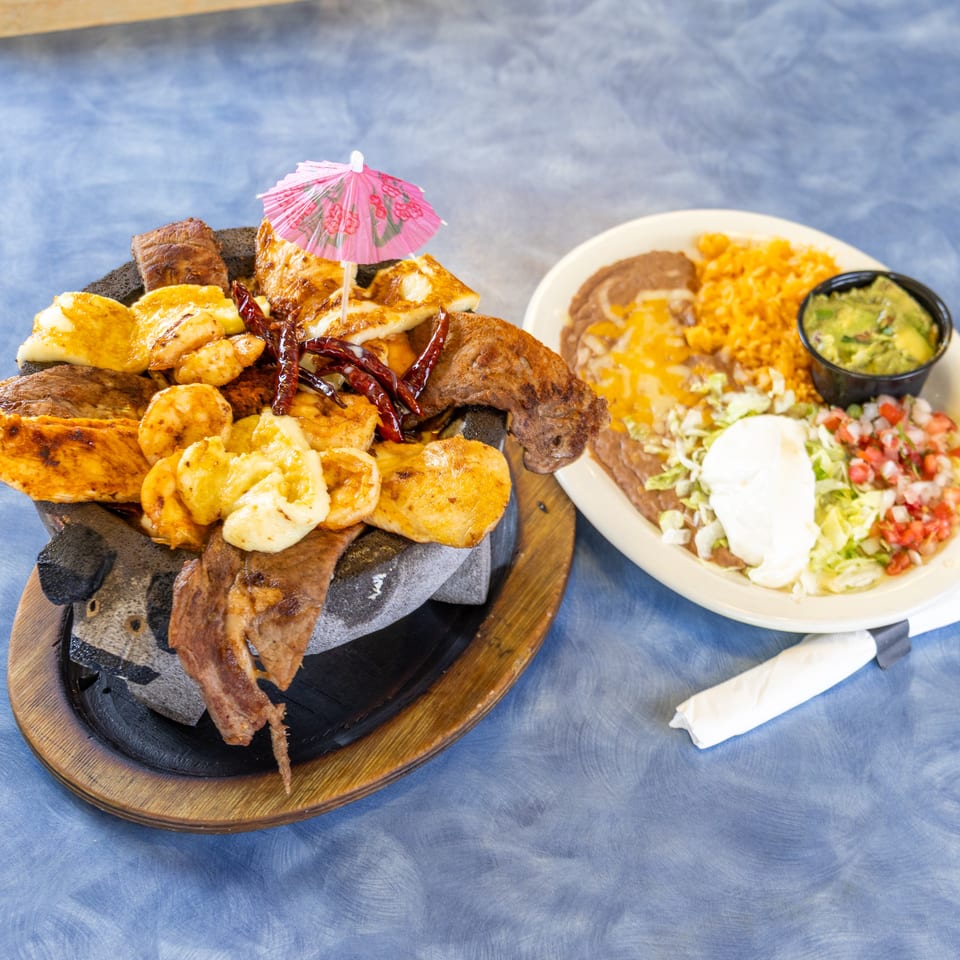 Grilled meats, shrimp, plantains, and cheese on a sizzling platter beside rice, beans, guacamole, salad, and salsa.