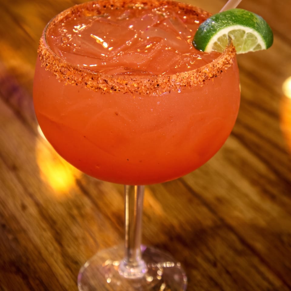 A vibrant red margarita with a chili-lime rim, ice, a straw, and a lime wedge on a wooden table.